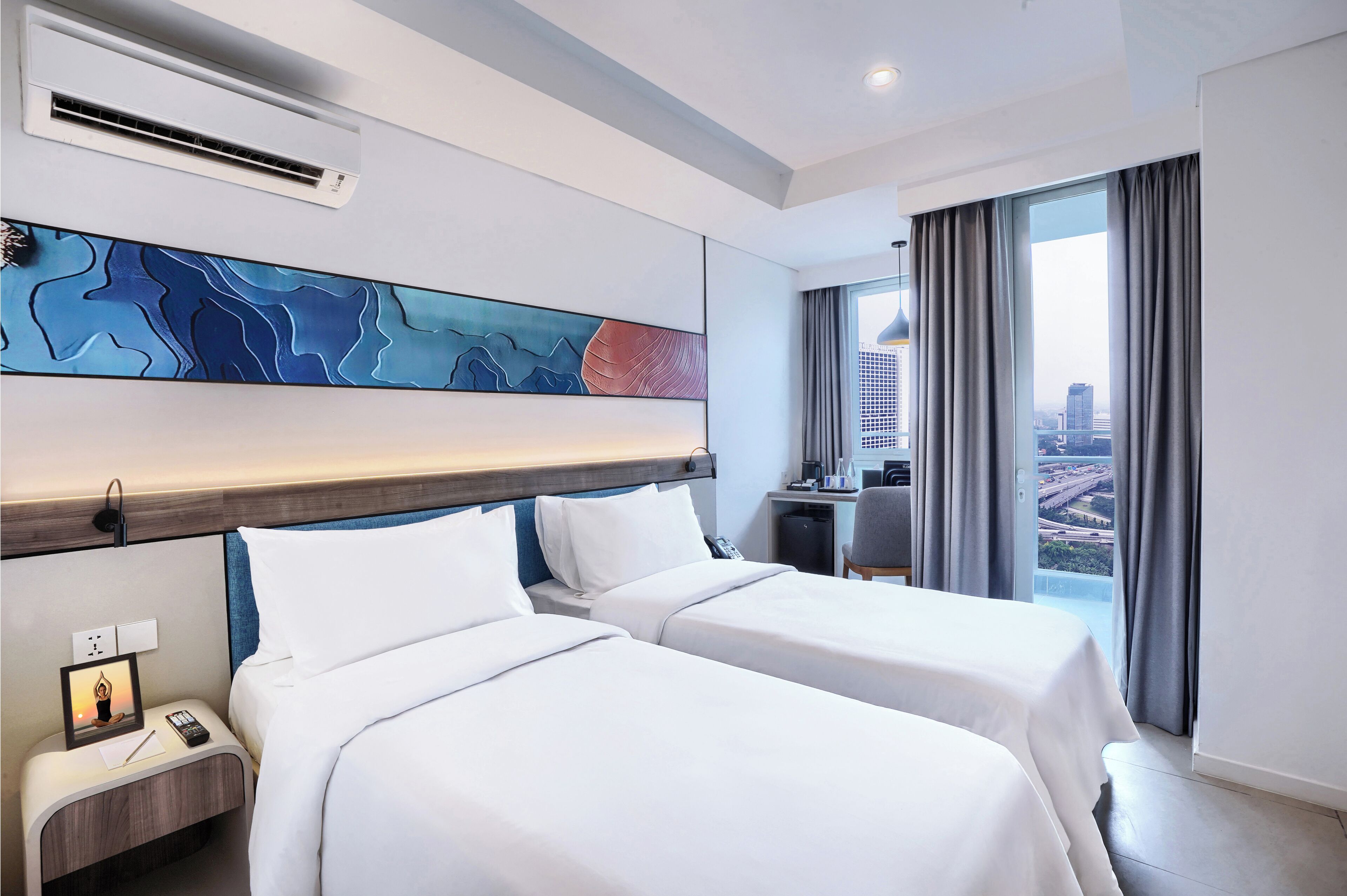 A hotel room with two beds, a city view, a modern abstract painting, and a desk with a chair.