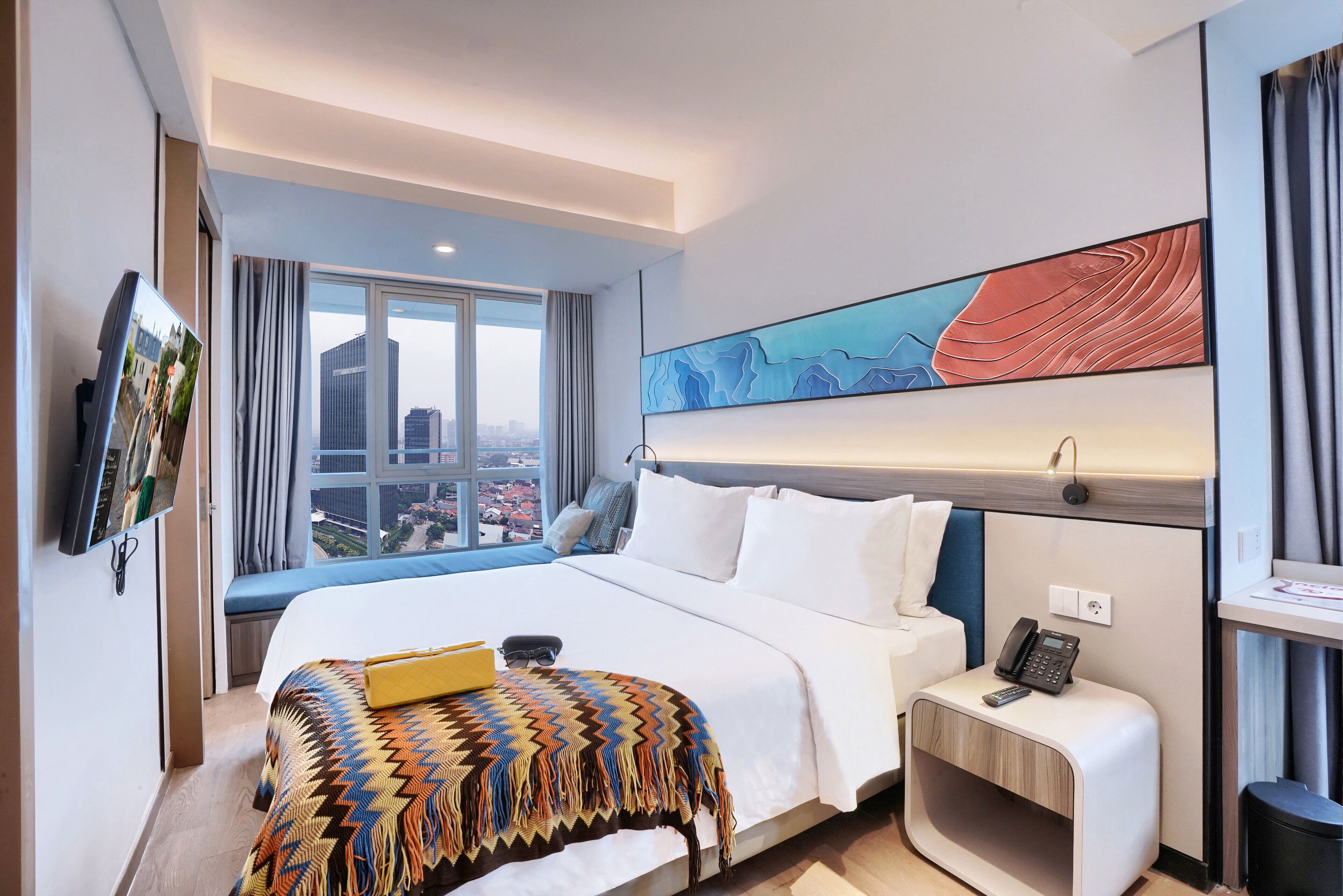 Een moderne hotelkamer met een groot bed, een nachtkastje met een telefoon, een aan de muur bevestigde televisie en uitzicht over de stad.