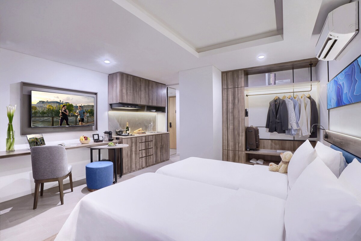 A modern hotel room with a large bed, a desk with a laptop, a television, and a wardrobe with hanging clothes.