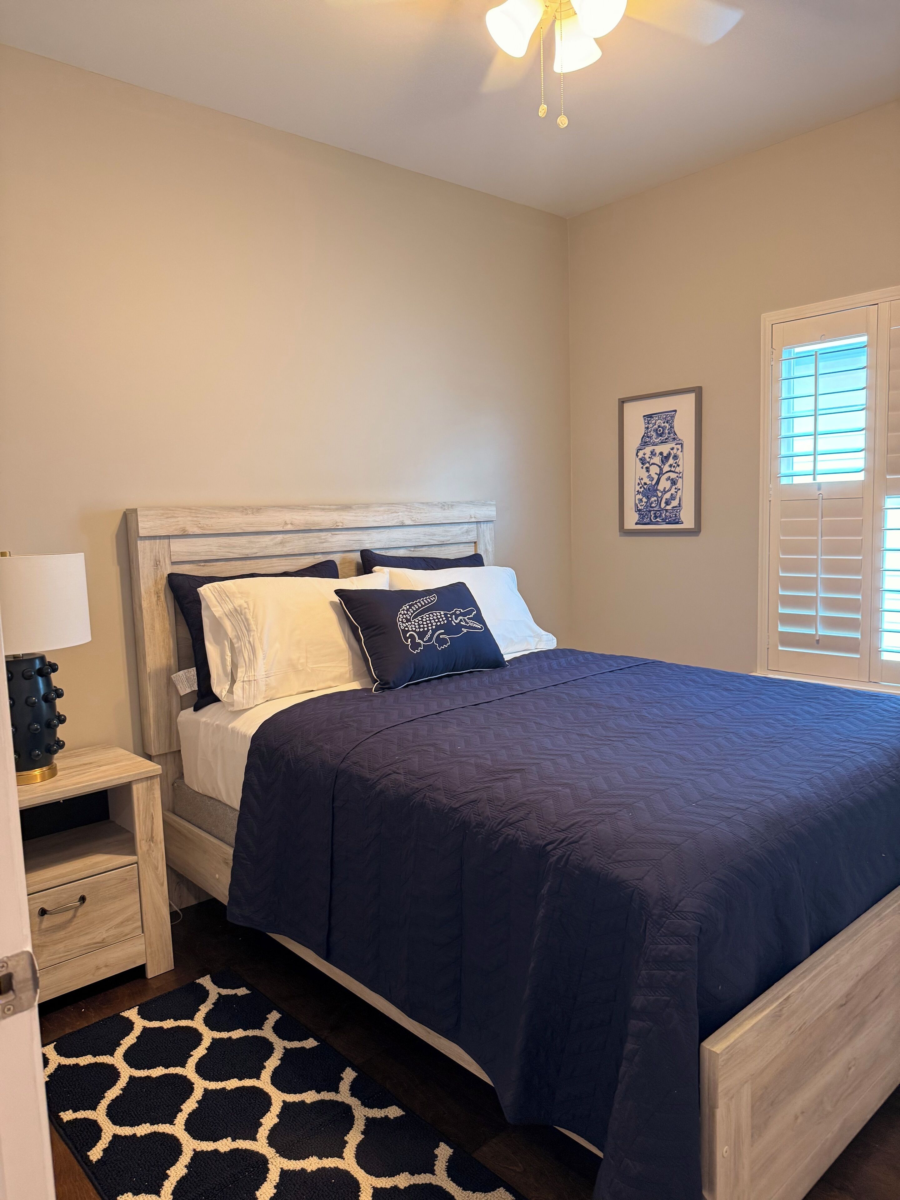 Quarto com cama, mesa de cabeceira, abajur, quadro emoldurado e janela com persianas.