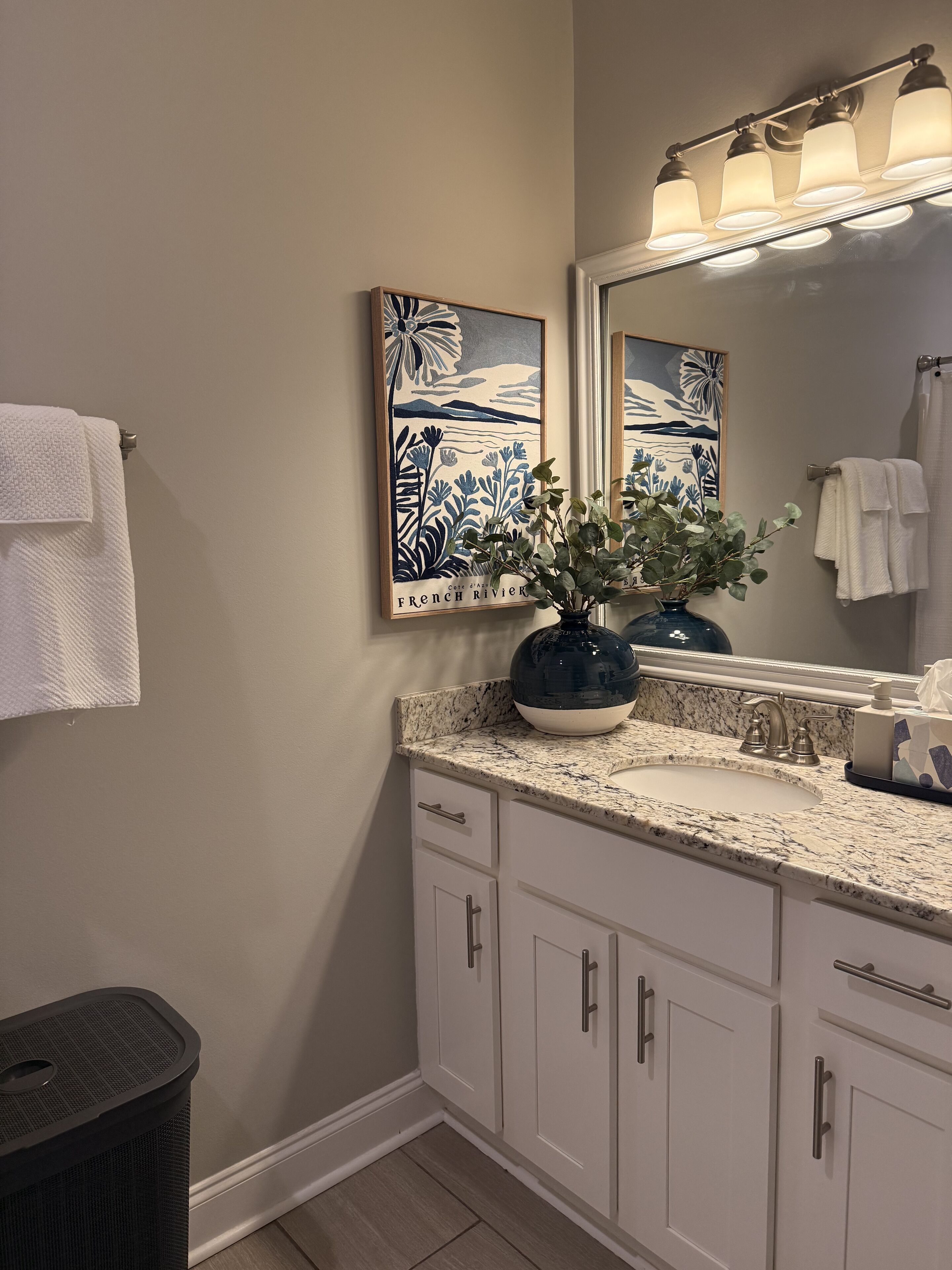 Une salle de bain avec un meuble-lavabo blanc, un miroir, un évier et une poubelle. Des serviettes sont accrochées au mur et un vase avec une plante est posé sur le meuble-lavabo.
