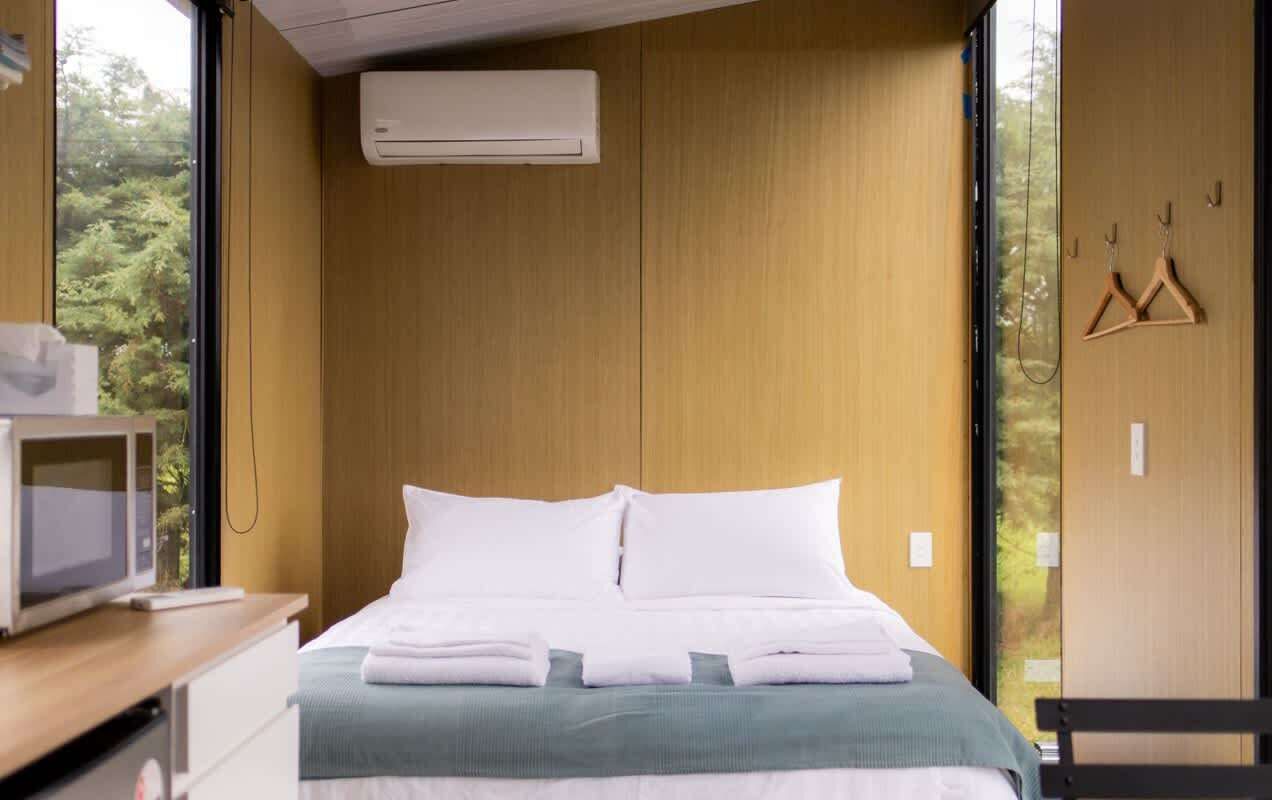 Una cama bien hecha con sábanas blancas, cabecera de madera y vista a árboles a través de un ventanal grande.