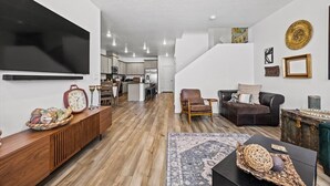 Ein geräumiges Wohnzimmer mit Holzboden, einer schwarzen Couch, einem braunen Ledersessel und einem Couchtisch. Ein großer Flachbildfernseher ist an der Wand befestigt, und im Hintergrund ist eine Küche zu sehen.