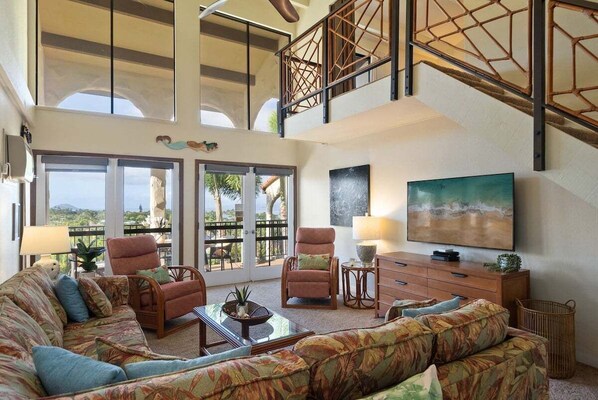 Amplia sala de estar con un sofá grande, dos sillones, una mesa de centro y una televisión. La habitación tiene un techo alto con vigas a la vista y un balcón con puertas de cristal.