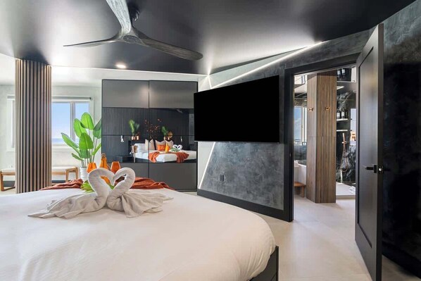 A modern hotel room with a large bed, a flat-screen TV mounted on the wall, and a view of the bathroom and living area.
