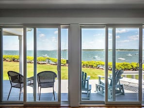 Una habitación con un gran ventanal corredera que da a una terraza exterior con sillas y una mesa, con vistas a un cuerpo de agua con barcos y vegetación.