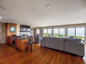 Un salón amplio con un sofá grande de color gris, piso de madera, una cocina con estufa a gas y vista al océano a través de amplias ventanas.
