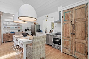 Cozinha moderna com armários brancos, sala de jantar com cadeiras de vime e uma grande porta de madeira.