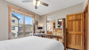 A bedroom with a large bed, wooden furniture, a ceiling fan, and a view of neighboring buildings. - Walk to Main Street | Hot Tub, Gym and Game Room (Breckenridge)