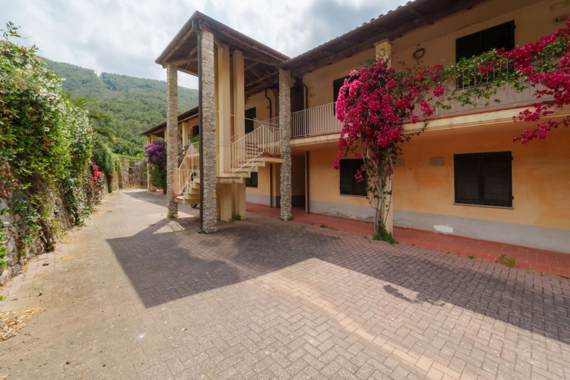 A building with a stone facade, a balcony with pink flowers, and a paved courtyard.