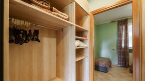 A wooden closet with shelves and hanging space, a bed with a patterned bedspread, and a view of a room with a curtain.