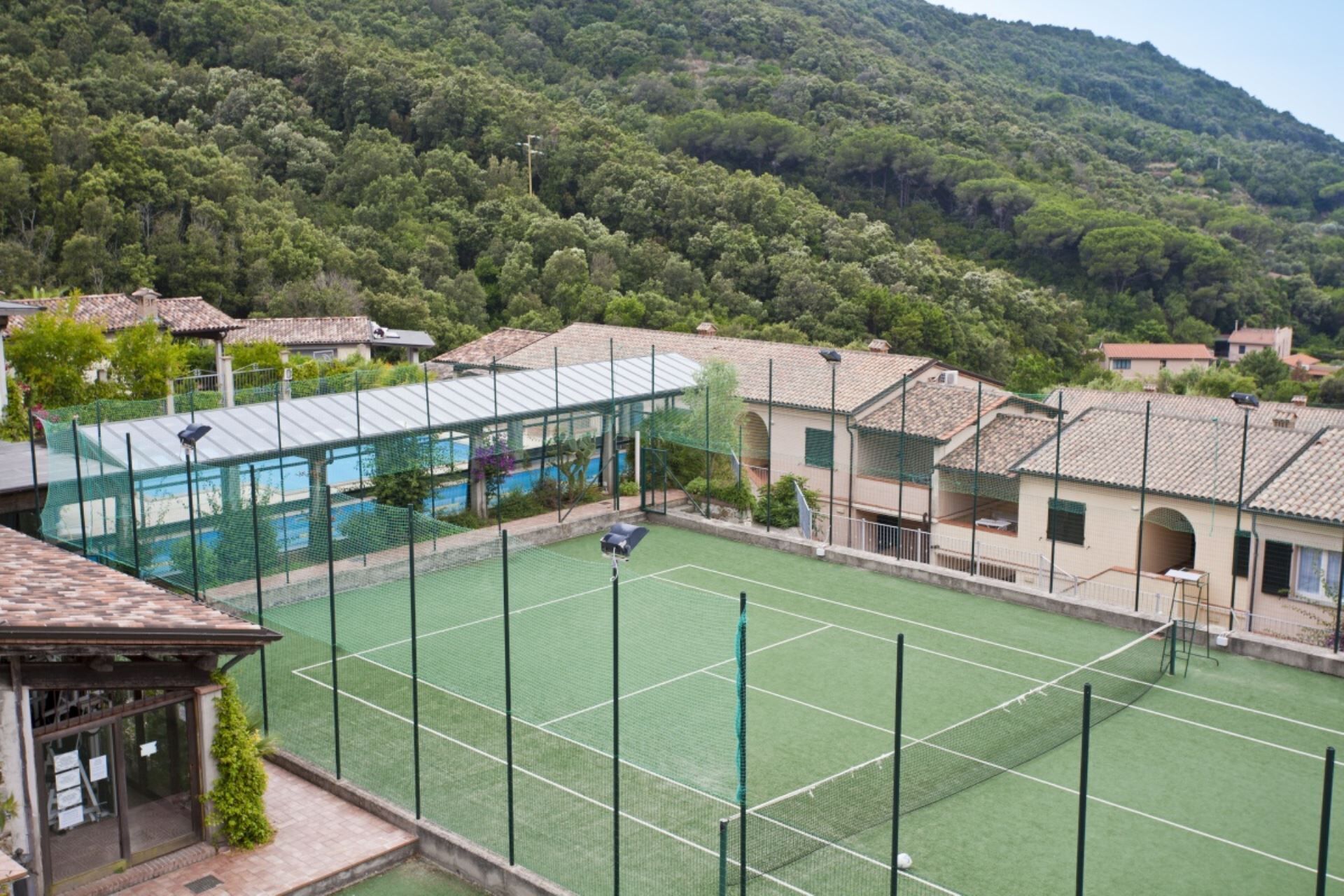 Un court de tennis avec un filet, entouré de bâtiments et avec une colline boisée en arrière-plan.