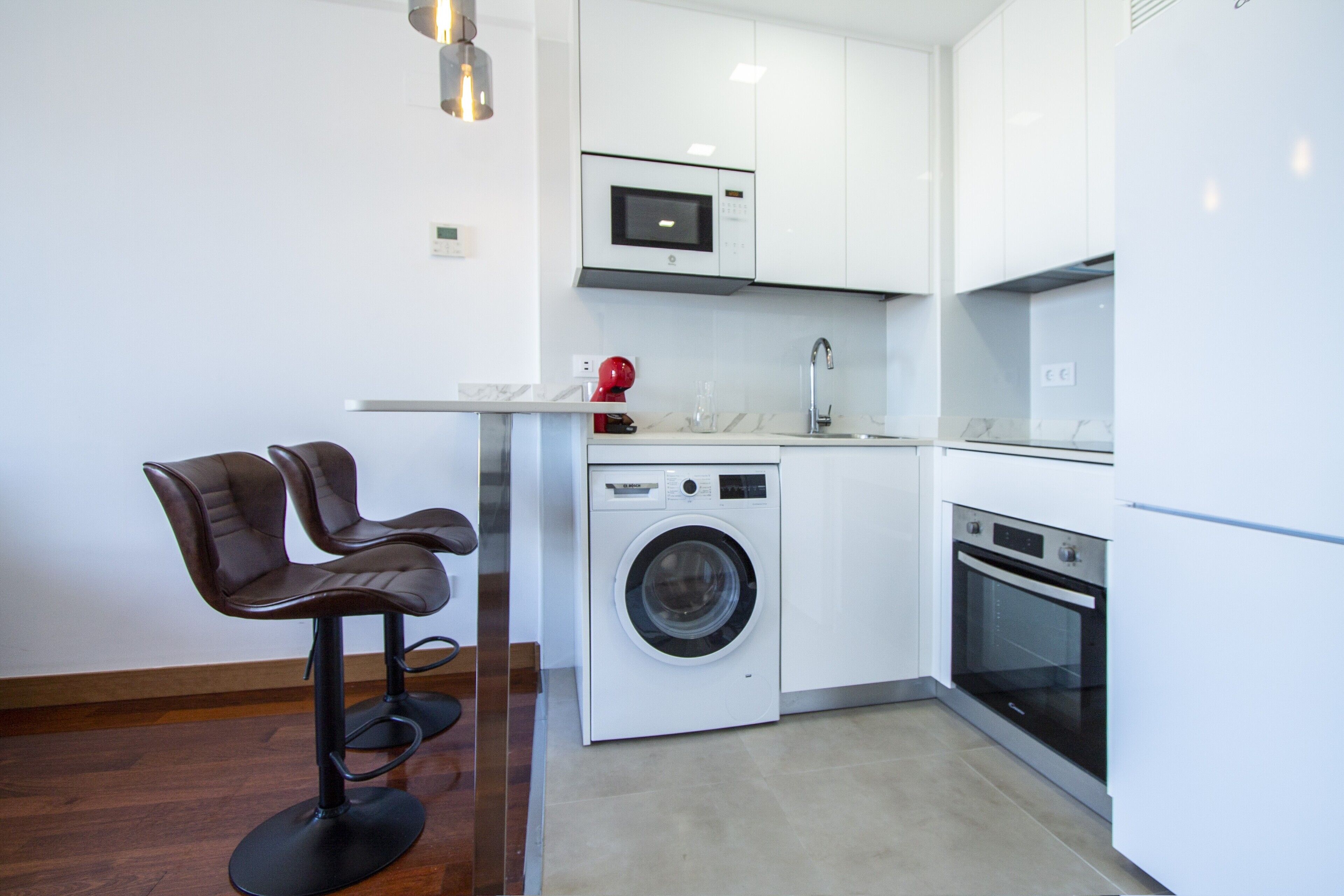 A compact kitchen with a washing machine, a microwave, and a refrigerator. There are two bar stools and a small countertop.
