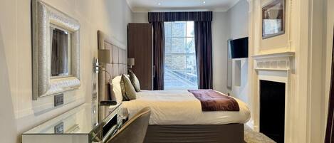 A hotel room with a bed, a desk, a chair, a fireplace, and a large window with curtains.