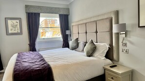 A neatly made bed with a headboard, a bedside table with a lamp, and a window with curtains.