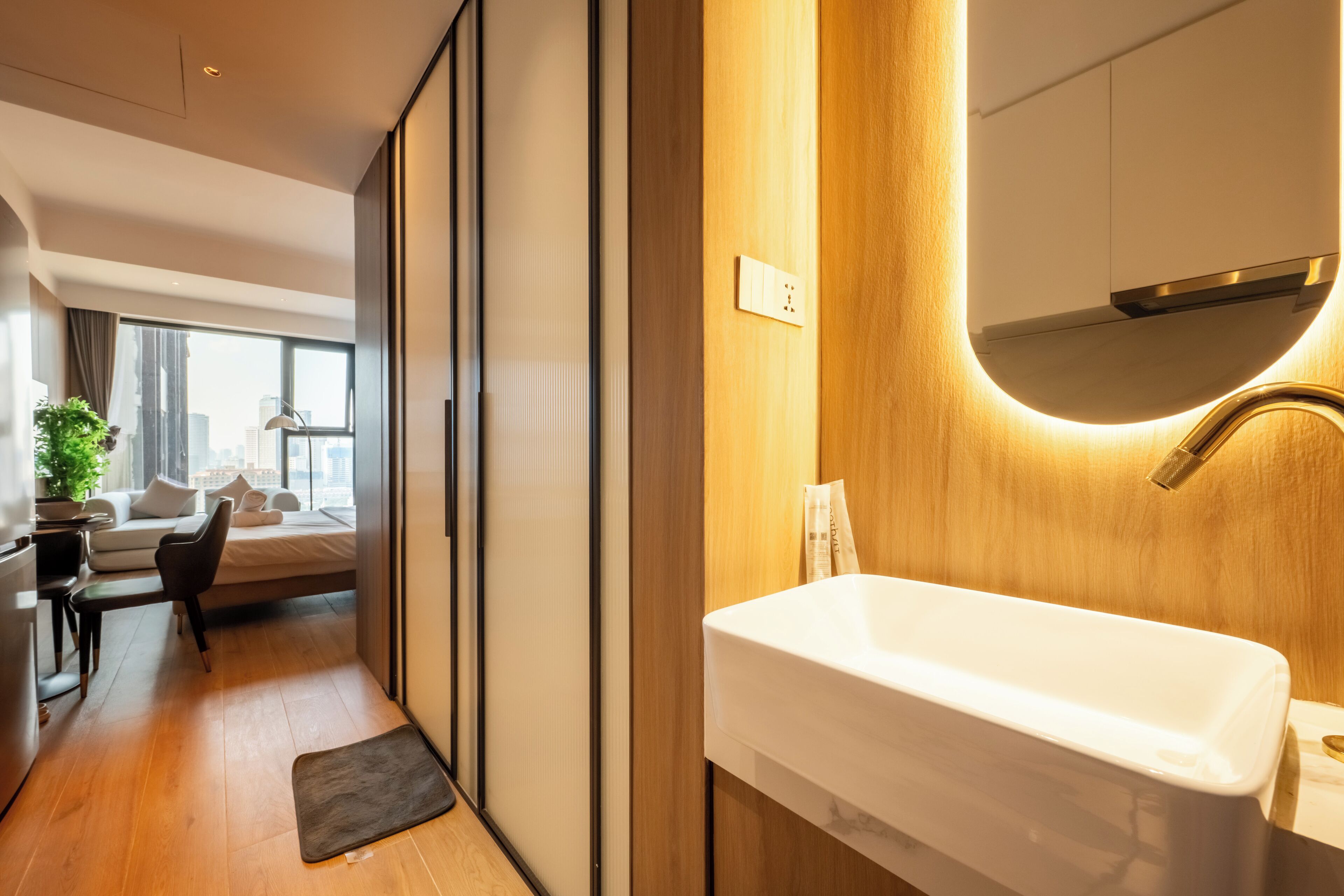 Una habitación de hotel moderna con piso de madera, un lavamanos blanco, un espejo grande y vista a la ciudad por la ventana.
