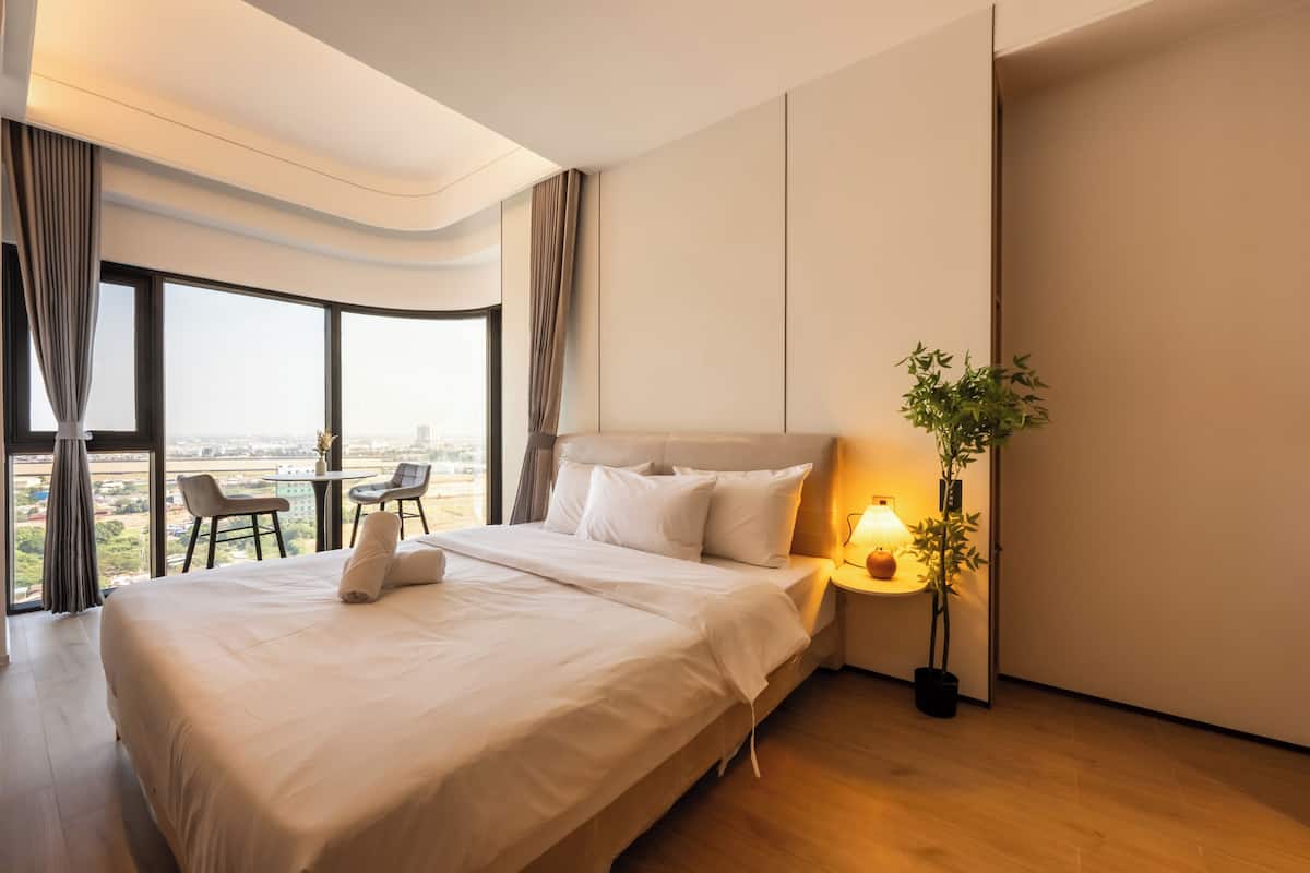 Ein modernes Hotelzimmer mit einem großen Bett, einem Nachttisch mit Lampe, einer Topfpflanze und Blick auf die Stadt durch große Fenster.