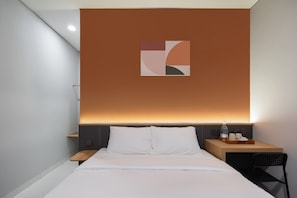 A hotel room with a bed, a nightstand with a lamp, a small table with a tray, and a wall-mounted artwork. - Cove Vaia (Bandung)