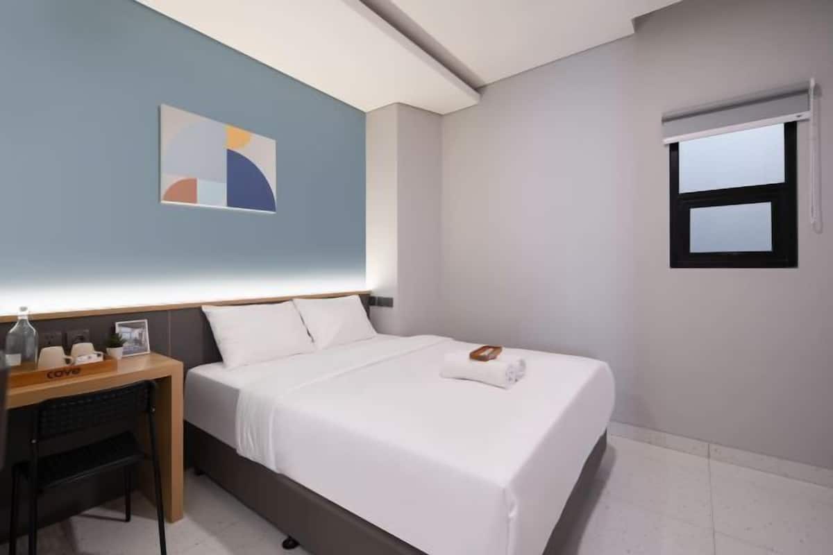 A hotel room with a bed, a desk, a chair, a window, and a wall-mounted artwork.
