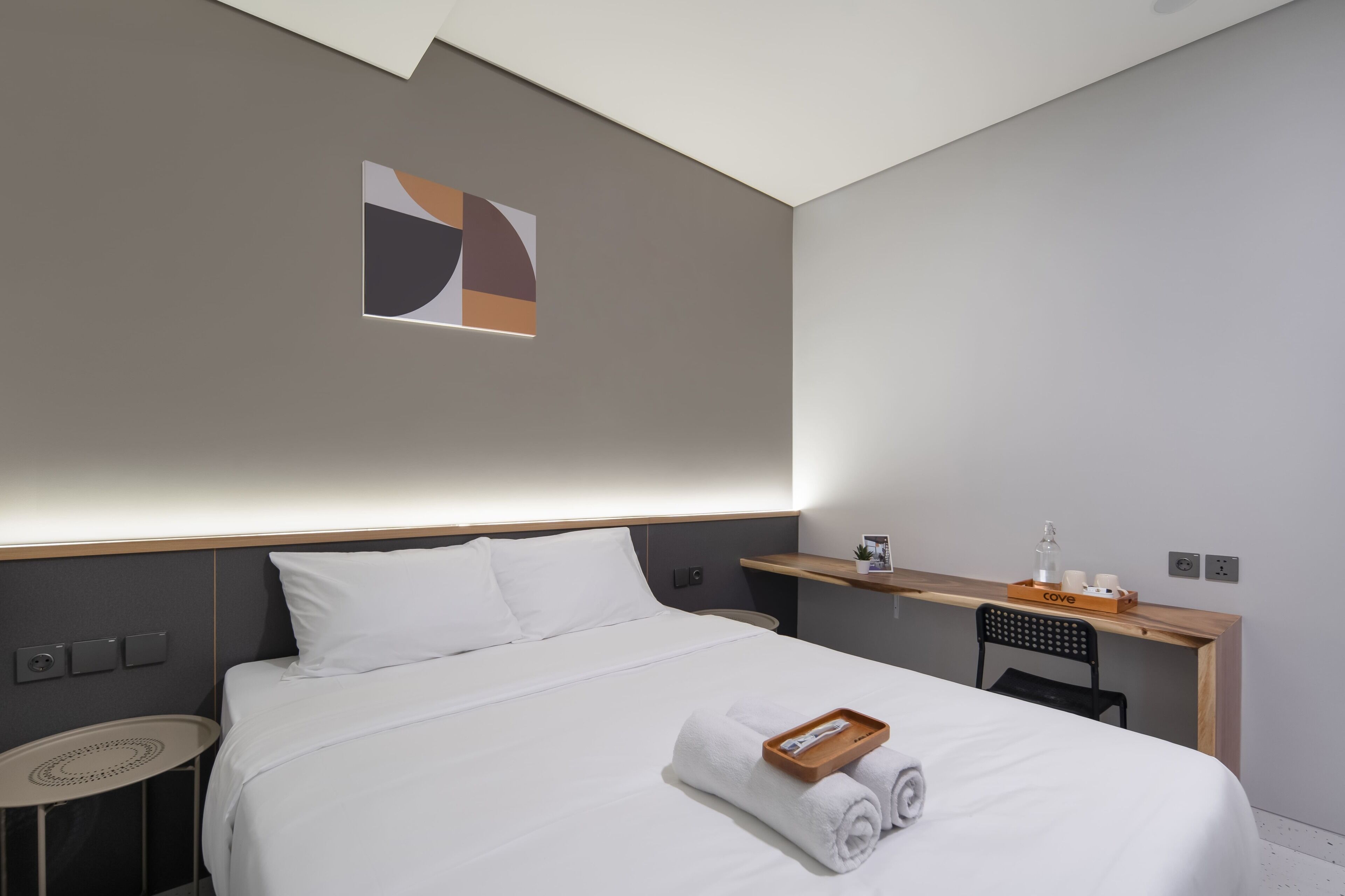 Una habitación de hotel moderna con una cama bien hecha, una mesita redonda pequeña, un escritorio con una silla y un estante fijado a la pared.