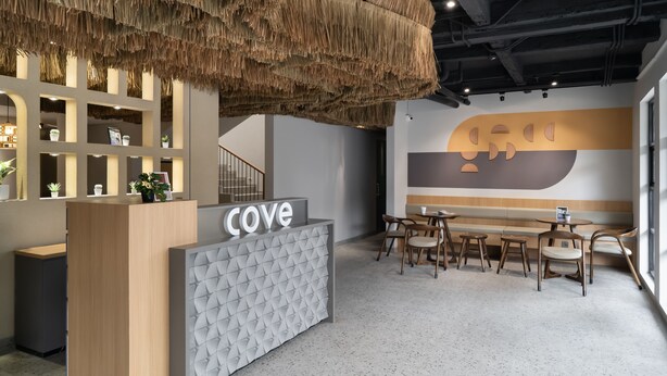 A reception desk with a 'Cove' sign, a hanging thatch ceiling, a staircase, and a wall with a geometric design. - Cove Vaia (Bandung)