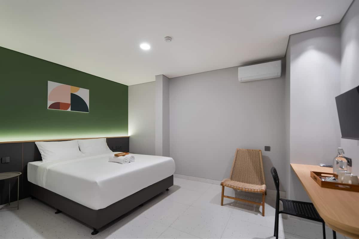 A modern hotel room with a bed, a chair, a desk, and a wall-mounted air conditioner.
