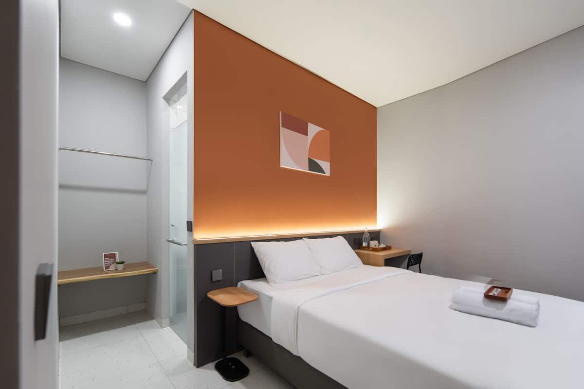 A modern hotel room with a bed, bedside tables, a nightstand, a wall-mounted artwork, and a small shelf with a plant.
