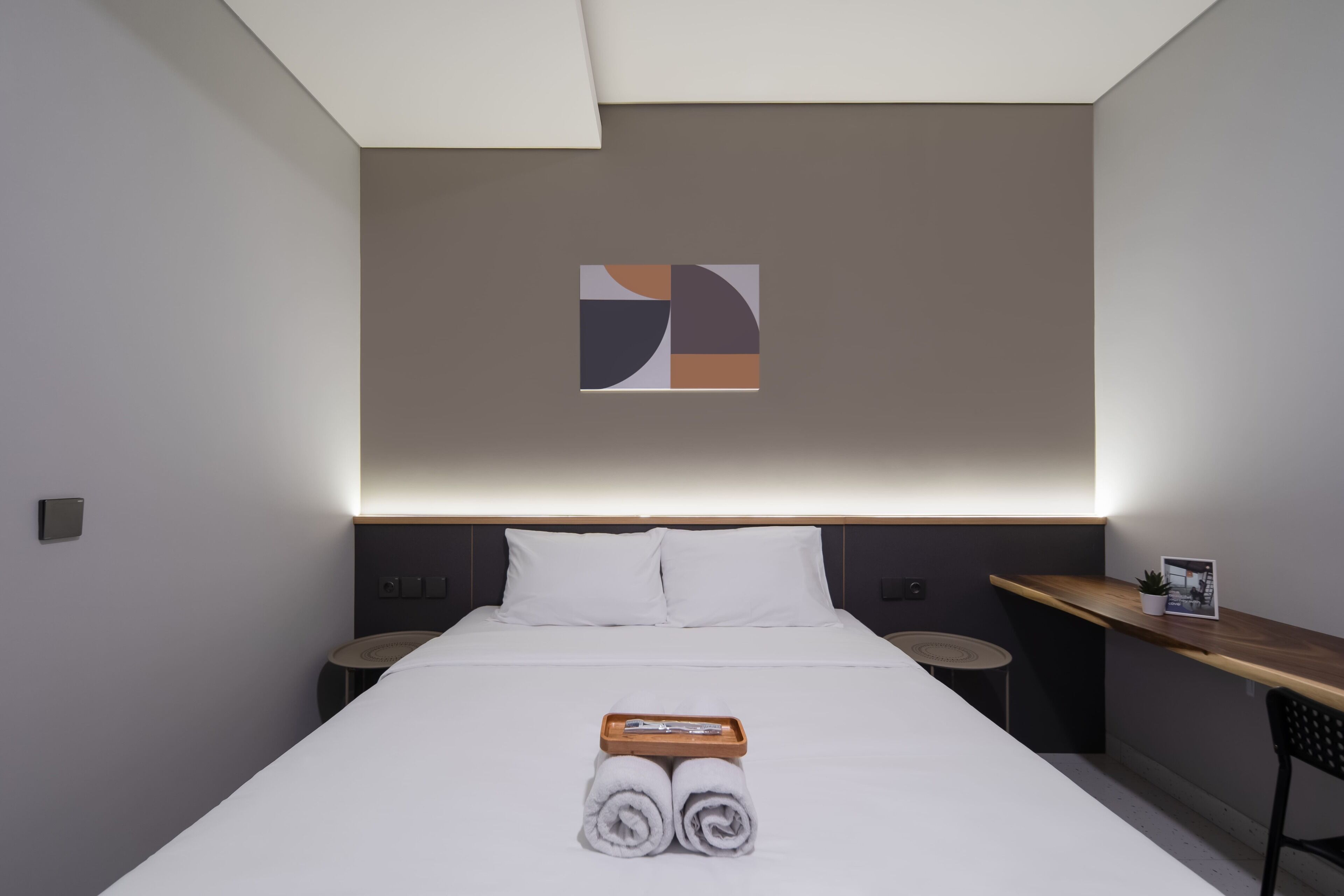 A modern hotel room with a neatly made bed, a bedside table with a plant, and a small desk area.