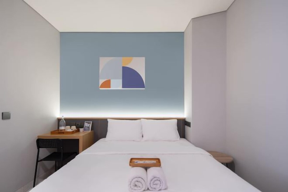 Une chambre d’hôtel moderne avec un lit soigneusement fait, une petite table d  ’appoint en bois, une chaise et une œuvre d’art abstraite accrochée au mur.