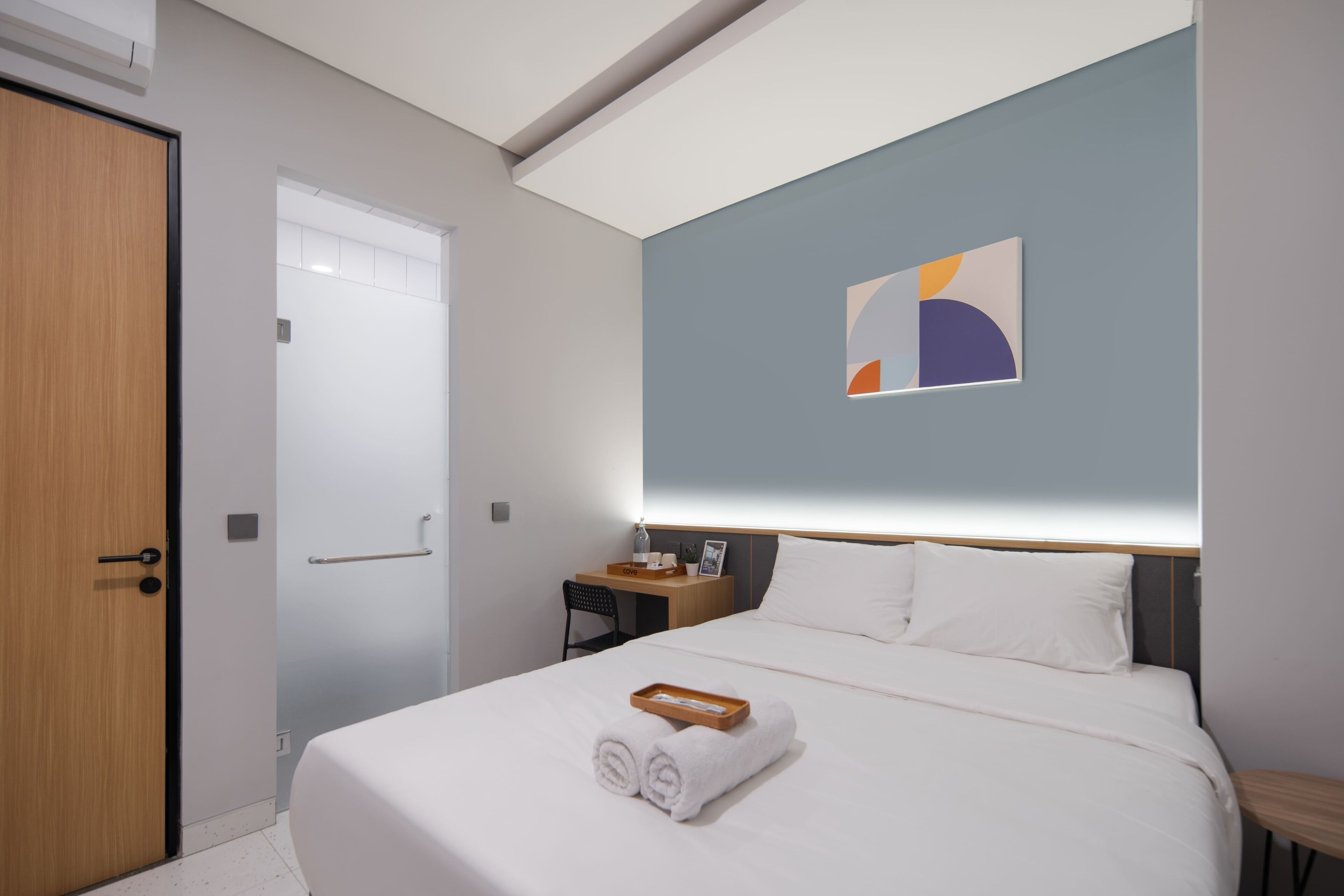 A hotel room with a bed, a desk, a chair, a bedside table, and a wall-mounted artwork.