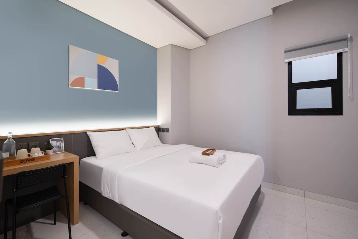 A neatly arranged hotel room with a bed, a desk, a chair, a small table with a bottle, a picture frame, and a wall-mounted artwork.