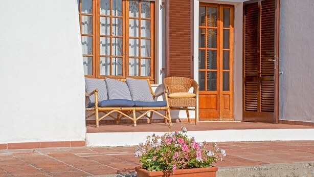 Un espace extérieur abrité, aménagé avec une chaise en osier et un banc rembourré, une porte en bois et un jardinière.