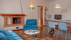 Un salon avec une cheminée en briques, des canapés bleus, une table basse ronde et un coin repas avec des chaises et un téléviseur.