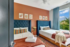 A hotel room with two beds, a ceiling fan, artwork on the wall, and a view of the outdoors.