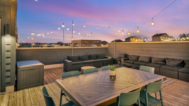 Een terras buiten met een houten tafel en stoelen, een bank en lichtslingers.
