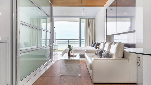 A modern living room with a glass coffee table, a white sofa, and a view of the ocean through large windows. - Chill Beachfront Vibes with 3BR Ocean Views & Pool (Sattahip)