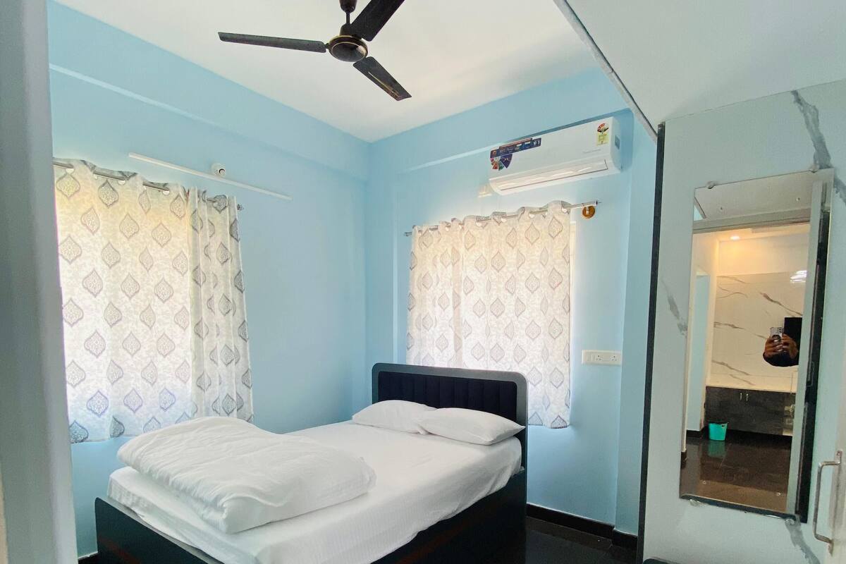 A bedroom with a bed, a ceiling fan, a window with curtains, a mirror, and a small bathroom with a sink and towel.