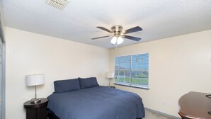 A bedroom with a bed, a ceiling fan, a window with blinds, and a nightstand with a lamp. - The Island Gem: Coastal Getaway (Corpus Christi)