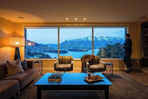 Una habitación con un ventanal grande, un sofá, dos sillones, una mesa de centro con piezas de ajedrez y aperitivos, y una persona de pie junto a la ventana.