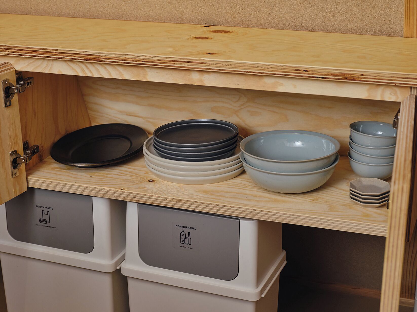 A kitchen cabinet with a wooden top shelf holding stacked plates and bowls, and two white trash bins below.
