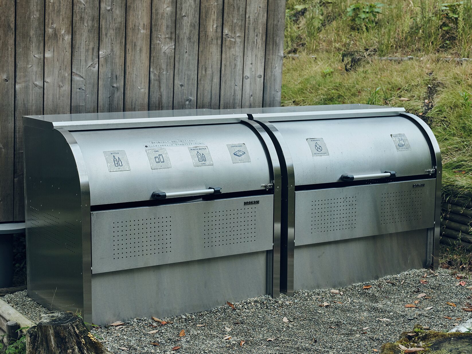 Un contenidor de metall per al reciclatge, amb compartiments etiquetats per a envasos de vidre, llaunes i amples de plàstic, col·locat a l'exterior amb una tanca de fusta al fons.