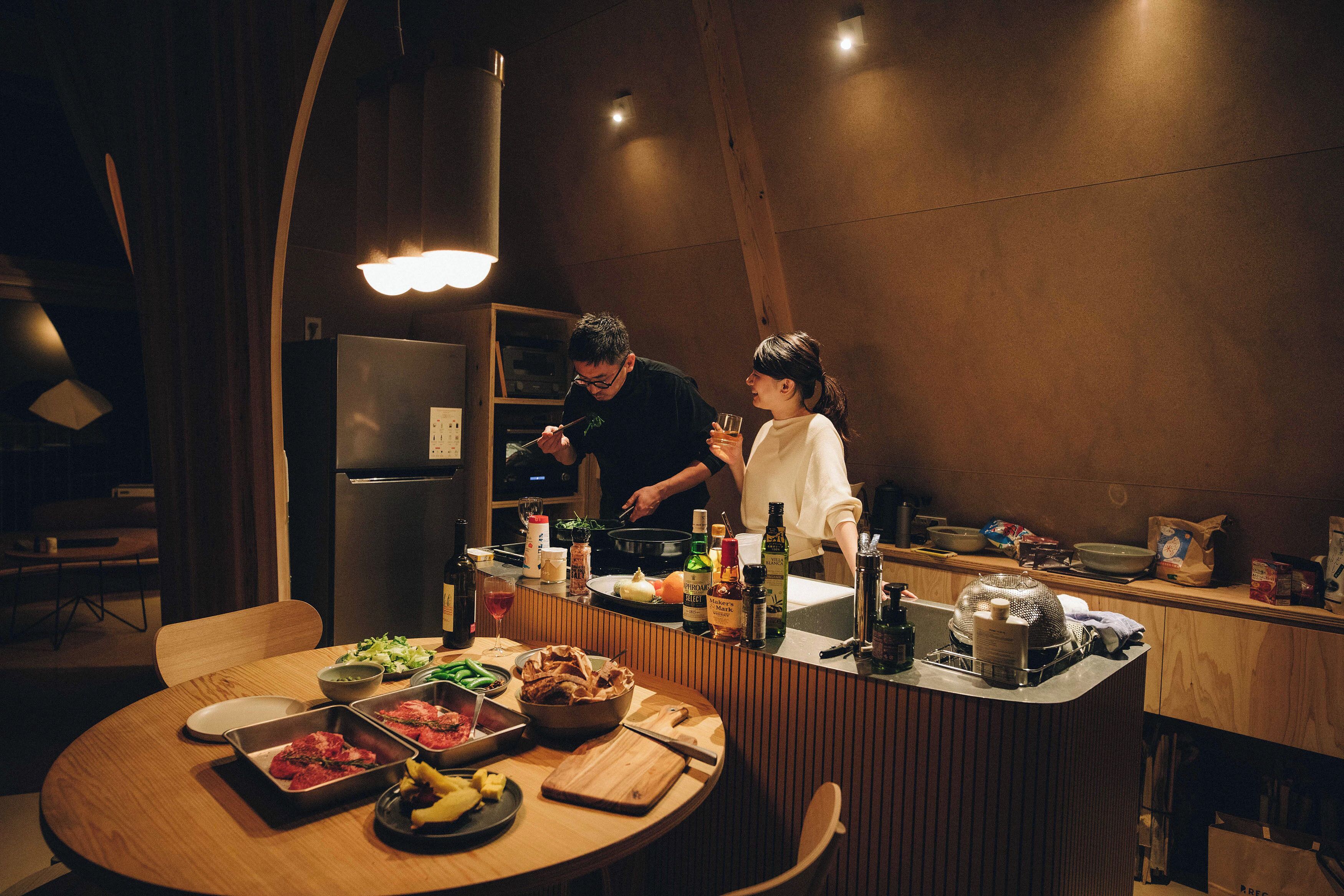 A kitchen with a wooden table set for a meal, a refrigerator, and various cooking utensils and ingredients.