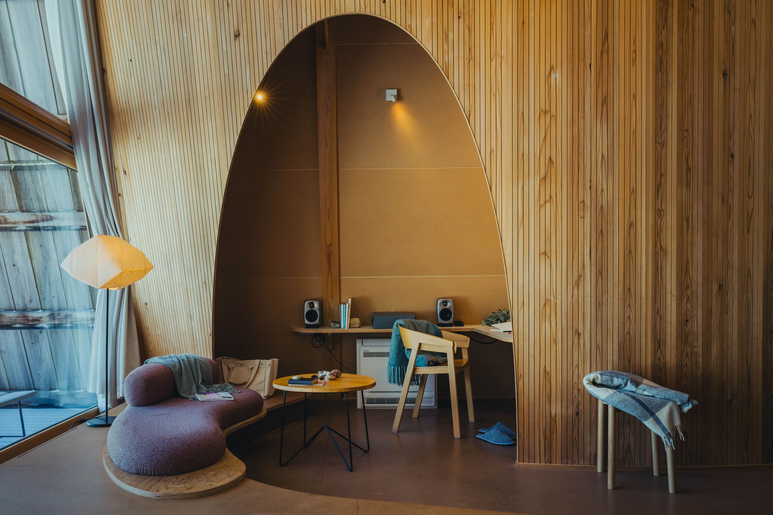 Ein gemütliches Zimmer mit einer gewölbten Holzwand, einem runden Tisch, einer gepolsterten Sitzecke und einem Schreibtisch mit Lautsprechern.