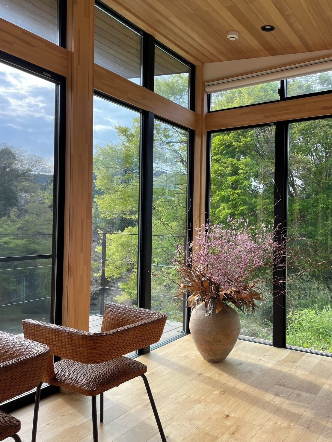 A spacious room with large windows offering a view of lush greenery. There is a wicker chair and a large vase with flowers on a wooden floor.