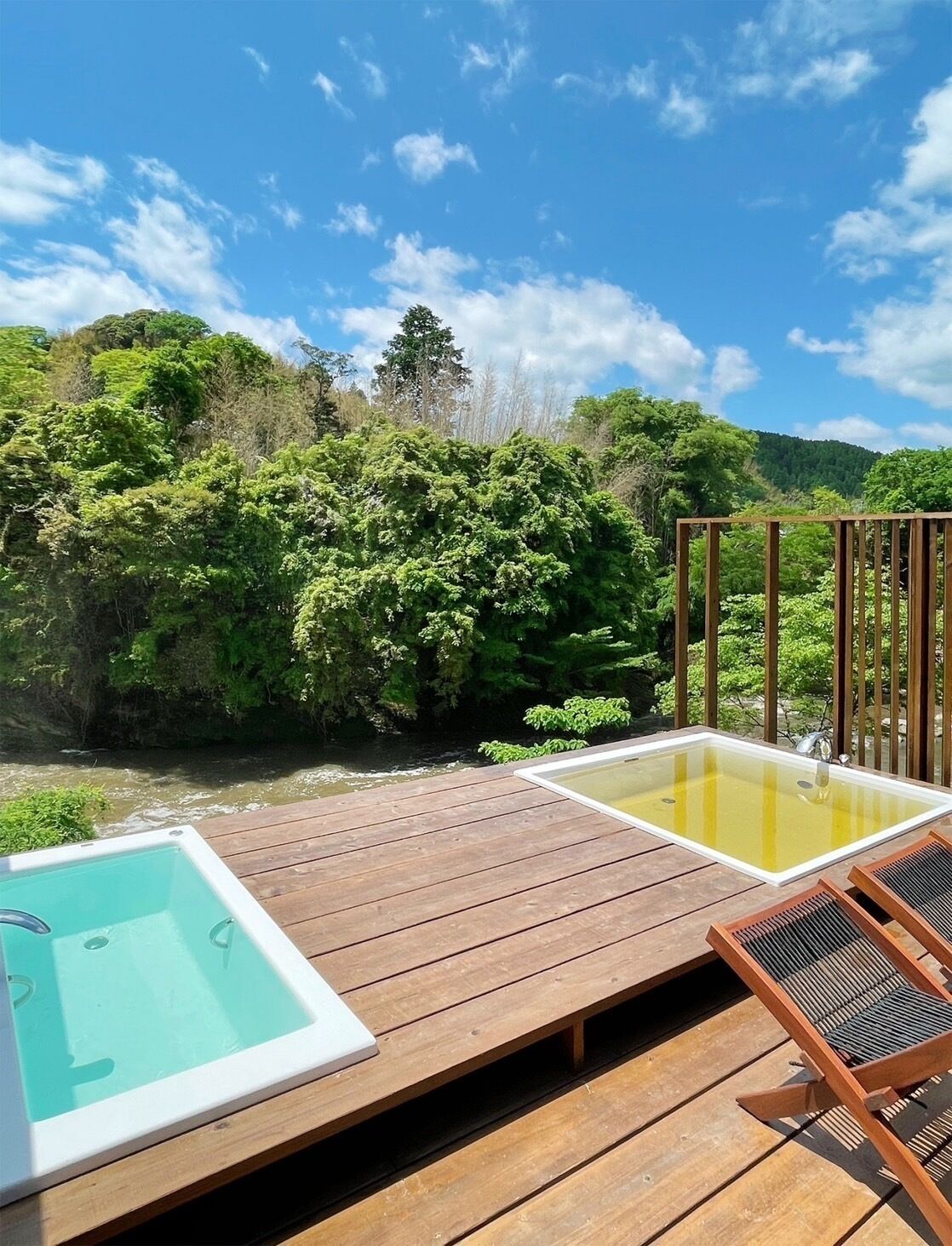 A wooden deck with a hot tub and two lounge chairs overlooking a river and forested hills.