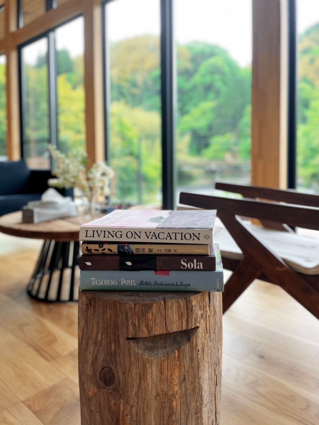 Una pila de llibres sobre un tamboret de fusta en una habitació amb grans finestres i vistes a la vegetació exterior.