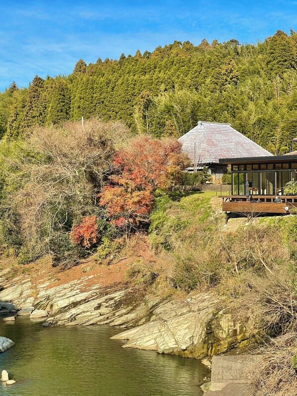 A house with a sloped roof, situated on a hillside with a view of a river and dense forest in the background.