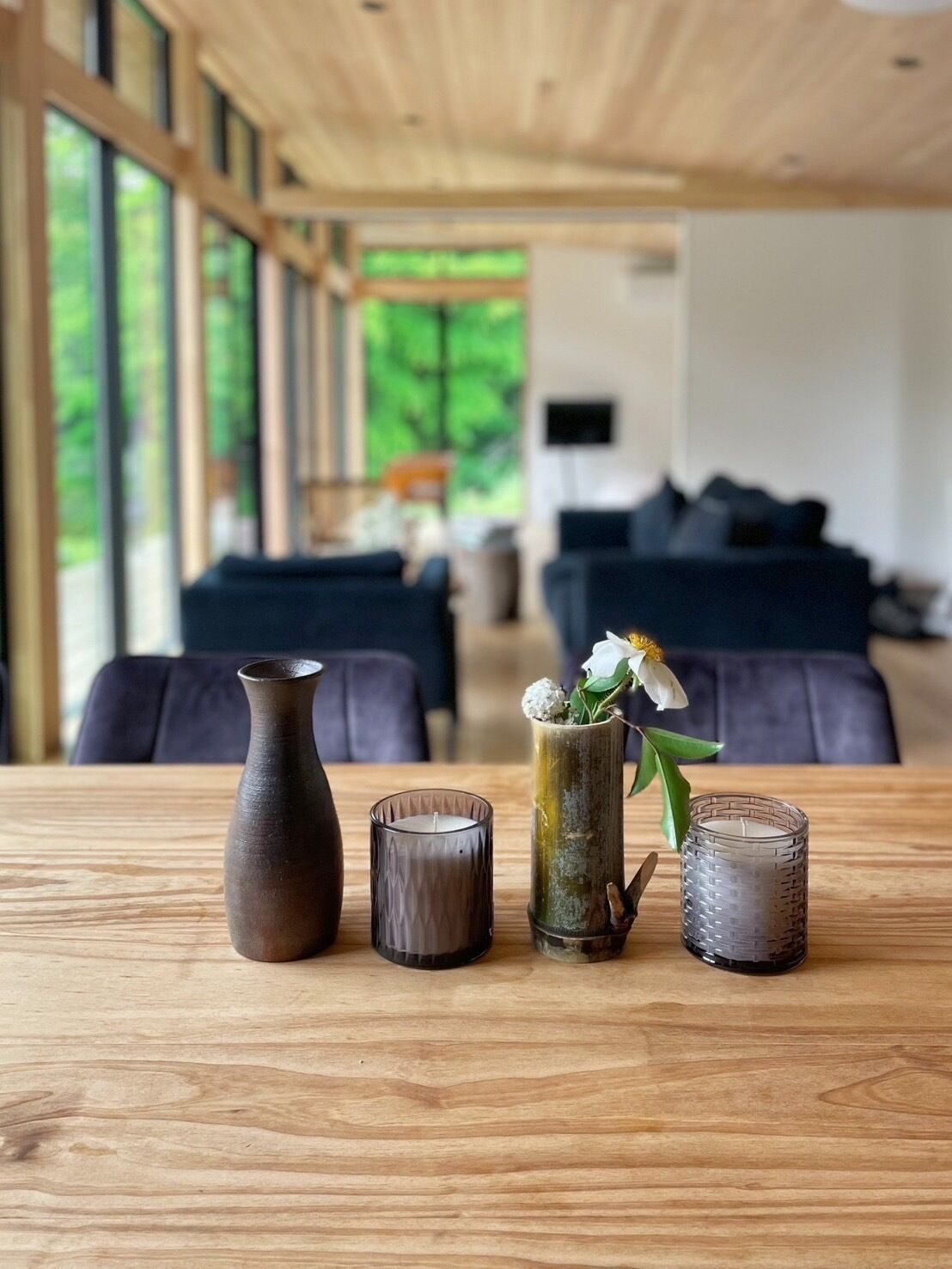 A wooden table with three vases and candles in a modern living room with large windows and a view of greenery outside.