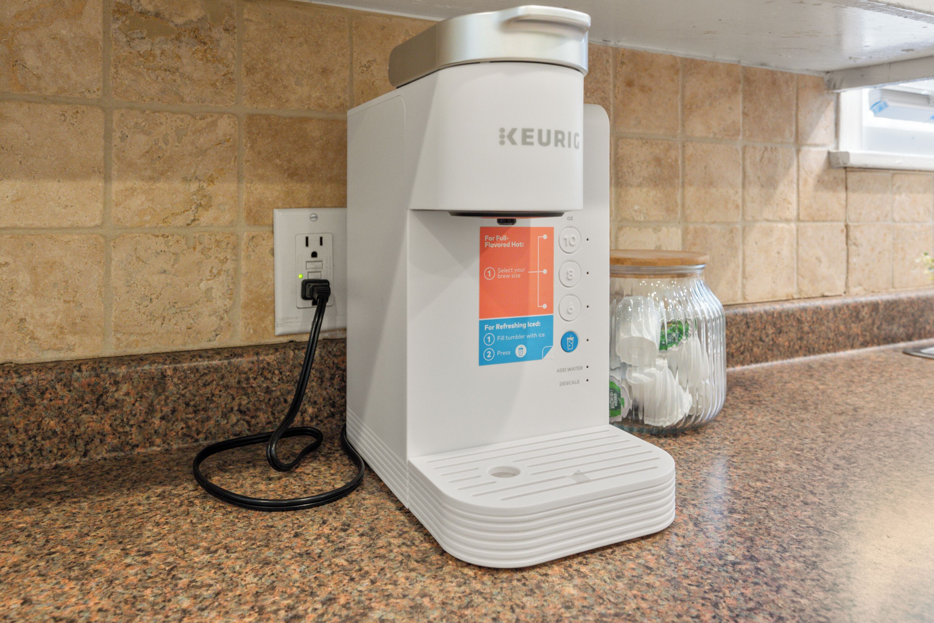 Una cafetera Keurig sobre una encimera de granito, con un tarro de vidrio con hielo y un enchufe de corriente al fondo.
