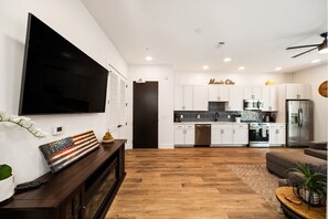 A modern kitchen with white cabinets, a stainless steel refrigerator, and a flat-screen TV mounted on the wall.